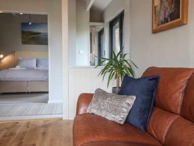 A room with a brown leather sofa with two cushions a potted plant near a window and a bedroom with a bed in the background at Sir George Baker Cabin in Uplyme