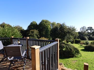 A wooden balcony with black railings two chairs and a table with wine and glasses overlooking a grassy garden with trees at Sir George Baker Cabin in Uplyme