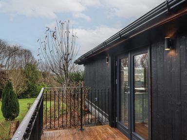 A balcony with wooden floor and black railing attached to a black wooden cabin with glass doors and outdoor lights overlooking a garden with trees at Sir George Baker Cabin in Uplyme