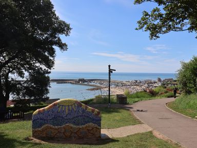 A coastal park with a mosaic bench overlooking a harbor and beach with people and boats at Sir George Baker Cabin in Uplyme