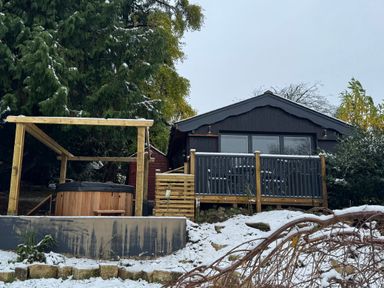 A cabin with a wooden deck and a hot tub under a wooden frame surrounded by snow and trees at Sir George Baker Cabin in Uplyme