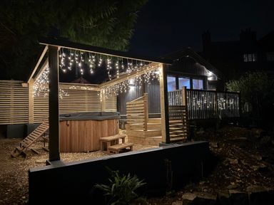 An outdoor wooden hot tub area with string lights a wooden lounge chair and a fenced deck attached to a house at Sir George Baker Cabin in Uplyme
