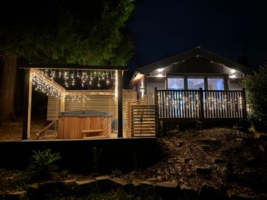 An outdoor patio with a hot tub under string lights next to a house with a balcony decorated with string lights at Sir George Baker Cabin in Uplyme