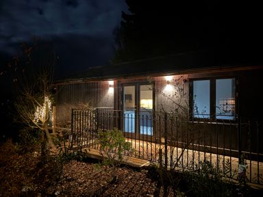 A cabin exterior at night with lit wall lights and a small fenced porch at Sir George Baker Cabin in Uplyme