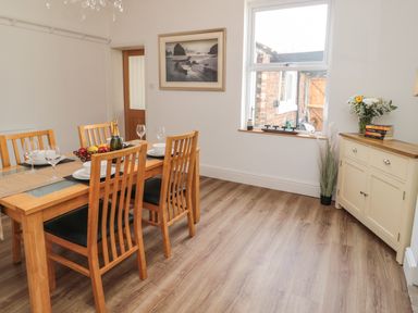 A dining room with a wooden table set with plates glasses and fruit a window a sideboard with flowers and books and a door at 18 Acklington Road in Amble