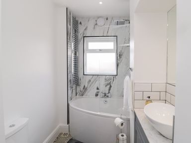 A bathroom with a corner bathtub shower combination a window above the tub a white toilet a sink on a counter and a towel rack at 18 Acklington Road in Amble