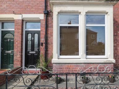 Front view of a brick house with a black door numbered 18 two white framed windows and several potted plants behind a decorative metal fence at 18 Acklington Road in Amble