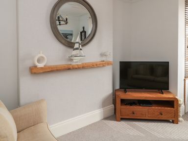 A corner of a living room with a wooden TV stand holding a television next to a beige armchair and a wall shelf with decorative items at 18 Acklington Road in Amble