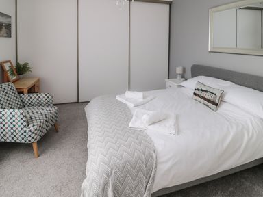 A bedroom with a bed with white linens and towels a patterned armchair a wooden table with a plant and framed picture and sliding door closets at 18 Acklington Road in Amble