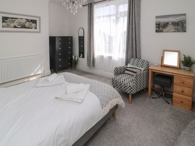A bedroom with a bed and folded towels a tall black chest of drawers a patterned armchair and a wooden desk with a mirror and plant at 18 Acklington Road Amble