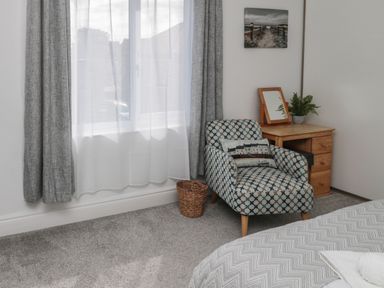 A bedroom corner with a patterned armchair, wooden desk with a mirror and plant, carpeted floor, and a window with curtains at 18 Acklington Road in Amble