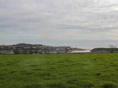 A grassy field with a village and body of water in the background with a cloudy sky at 18 Acklington Road in Amble