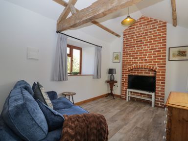 A living room with a blue sofa a television on a white stand in a brick fireplace alcove exposed wooden beams and a window with curtains at Keepers Barn Bromyard