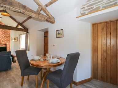 A dining area with a round wooden table set for two and dark upholstered chairs in a room with exposed wooden beams and a brick fireplace at Keepers Barn in Bromyard