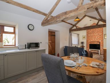 A kitchen and dining area with wooden beams, a table set for two, cabinets with a microwave and toaster, and a living room with a TV and brick fireplace at Keepers Barn Bromyard
