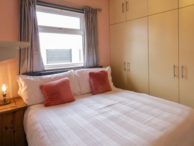 A bedroom with a bed with white and pink pillows a window with curtains a wooden bedside table with a lamp and built in wardrobes at Shore Road in Culdaff County Donegal