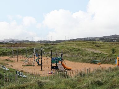 An outdoor playground with swings, climbing structures, and slides surrounded by grass and wooden fencing at Shore Road in Culdaff County Donegal