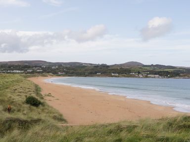 A sandy beach with grassy dunes leading to water and hills with houses in the distance at Shore Road in Culdaff County Donegal