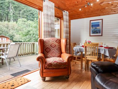 A wooden interior room with an armchair near a dining table set for four and a deck with outdoor chairs at Trout River Retreat Clifford Bridge near Moretonhampstead