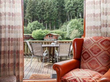 A seating area with an armchair and curtains leading to a wooden deck with a table and chairs overlooking trees and cabins at Trout River Retreat Clifford Bridge near Moretonhampstead