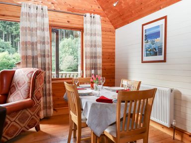 A dining table set for four next to a window with curtains and an armchair in a wooden room at Trout River Retreat Clifford Bridge near Moretonhampstead