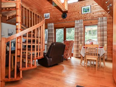 A wooden living room with a spiral staircase leather and patterned chairs and a dining table beside windows at Trout River Retreat Clifford Bridge near Moretonhampstead