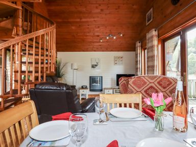 A dining table set with plates wine glasses and flowers in a wooden cabin with spiral staircase and seating area at Trout River Retreat Clifford Bridge near Moretonhampstead
