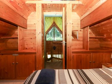 A bedroom with wooden walls and cabinets a bed with striped bedding and a doorway leading to a balcony with a window showing a forest view at Trout River Retreat in Clifford Bridge near Moretonhampstead