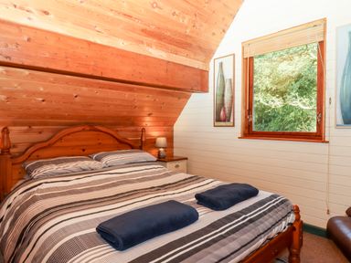 A bedroom with a wooden bed with striped bedding and two folded towels on the bed next to a window showing green trees at Trout River Retreat Clifford Bridge near Moretonhampstead