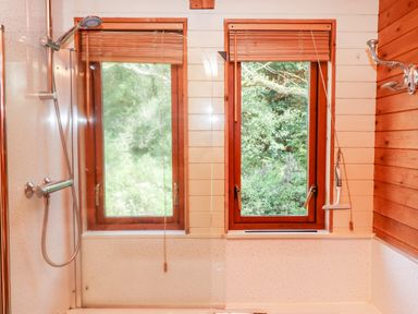 A bathroom with a showerhead bathtub two windows with wooden blinds and a cleaning squeegee at Trout River Retreat Clifford Bridge near Moretonhampstead