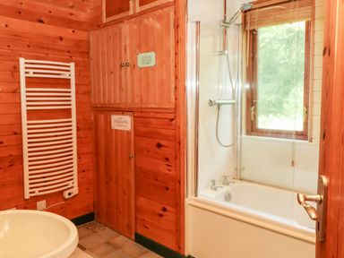 A bathroom with wooden walls a white bathtub with shower and a window above it a white sink and a white towel radiator at Trout River Retreat Clifford Bridge near Moretonhampstead