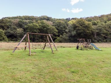 A playground with swings tire swing and slide on a grassy field with trees in the background at Trout River Retreat Clifford Bridge near Moretonhampstead