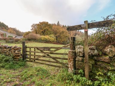 A wooden gate next to a stone wall and a signpost in a grassy field with trees and buildings in the background at Trout River Retreat in Clifford Bridge near Moretonhampstead