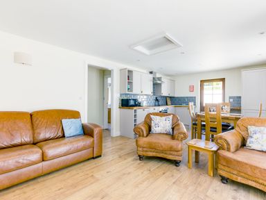 A living room with brown leather sofas and armchairs next to a kitchen with white cabinets and a wooden dining table at Chy Pol in St Minver
