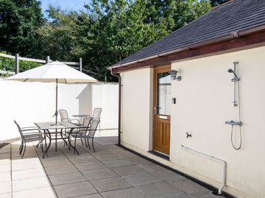 A patio with a table umbrella chairs and an outdoor shower attached to a building wall at Chy Pol in St Minver