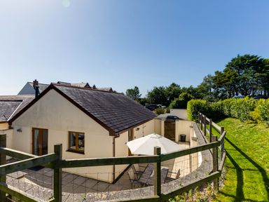 A small fenced outdoor patio area with a table umbrella and chairs next to a cream colored building with a tiled roof surrounded by grass and trees at Chy Pol in St Minver