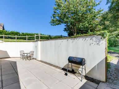 A patio area with a table and two chairs and a barbecue grill against a white wall at Chy Pol in St Minver