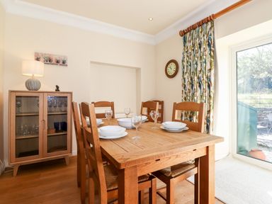 A dining room with a wooden table and chairs set with plates and glasses next to a glass cabinet and a window with patterned curtains at Silver How Ambleside