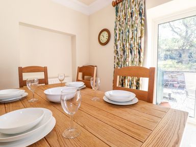 A dining room with a wooden table set with white plates and glasses wooden chairs a wall clock floral curtains and a glass door showing an outdoor area at Silver How in Ambleside
