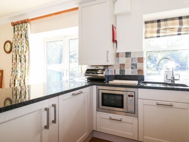 A kitchen area with white cabinets a built-in microwave black countertop a sink under a window and curtains beside glass doors at Silver How in Ambleside