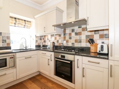A kitchen with white cabinets a black countertop a microwave oven a gas stove a toaster and a sink with a window and checked blinds at Silver How Ambleside