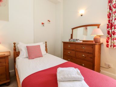 A bedroom with a single wooden bed with white and red bedding a wooden chest of drawers with a lamp and a mirror and floral curtains at Silver How in Ambleside
