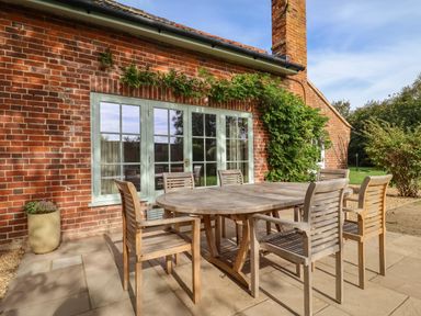 An outdoor patio with a wooden table and six chairs next to a red brick house at Rookery Cottage in Kelsale near Saxmundham