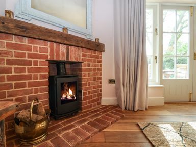 A brick fireplace with a wood burning stove and a brass container with firewood next to a window with curtains and a wooden floor at Rookery Cottage in Kelsale near Saxmundham