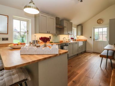 A kitchen with wooden countertops gray cabinets a dining bench and table and a door to the outside at Rookery Cottage in Kelsale near Saxmundham