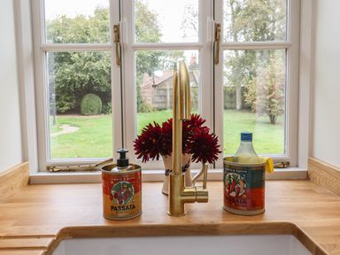 A wooden kitchen countertop with a brass faucet a flower vase and tomato passata containers near a window with a garden view at Rookery Cottage in Kelsale near Saxmundham