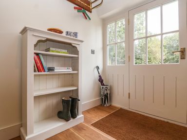A white bookshelf with books and boots next to a door with glass panels and an umbrella stand at Rookery Cottage in Kelsale near Saxmundham