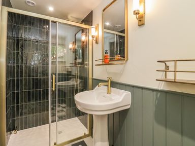 A bathroom with a white pedestal sink with a gold faucet a gold framed mirror two gold wall lights a glass shower enclosure with black vertical tiles and a gold frame