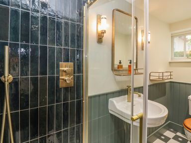 A bathroom with a shower area with dark tiles a white sink with a brass faucet a mirror and a window at Rookery Cottage in Kelsale near Saxmundham