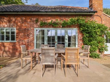 An outdoor patio with a wooden dining table and six chairs in front of a brick house with green framed windows and doors at Rookery Cottage in Kelsale near Saxmundham